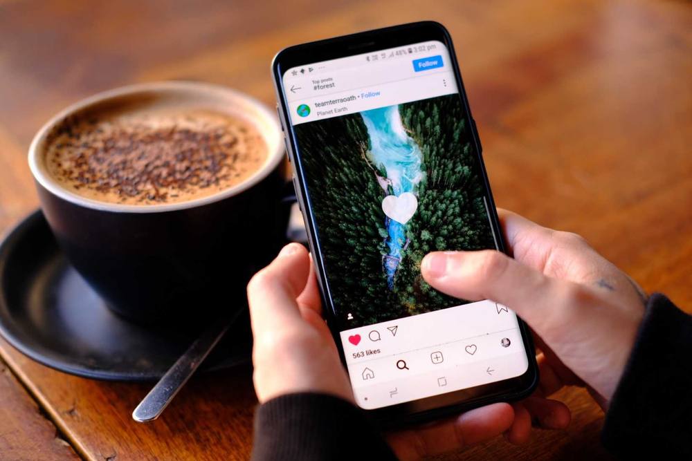 Pessoa rolando o feed do Instagram no celular, ao lado de um café